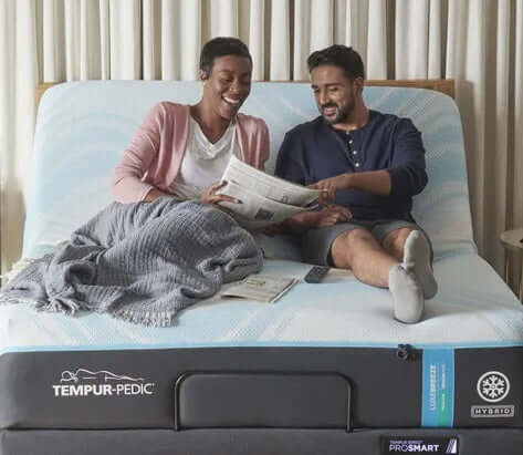 Couple enjoying comfort on a Tempur-Pedic hybrid mattress while reading a newspaper together in a cozy bedroom setting.
