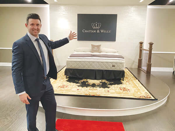 A smiling man in a suit proudly showcases a Chattam & Wells mattress in a stylish showroom setting.
