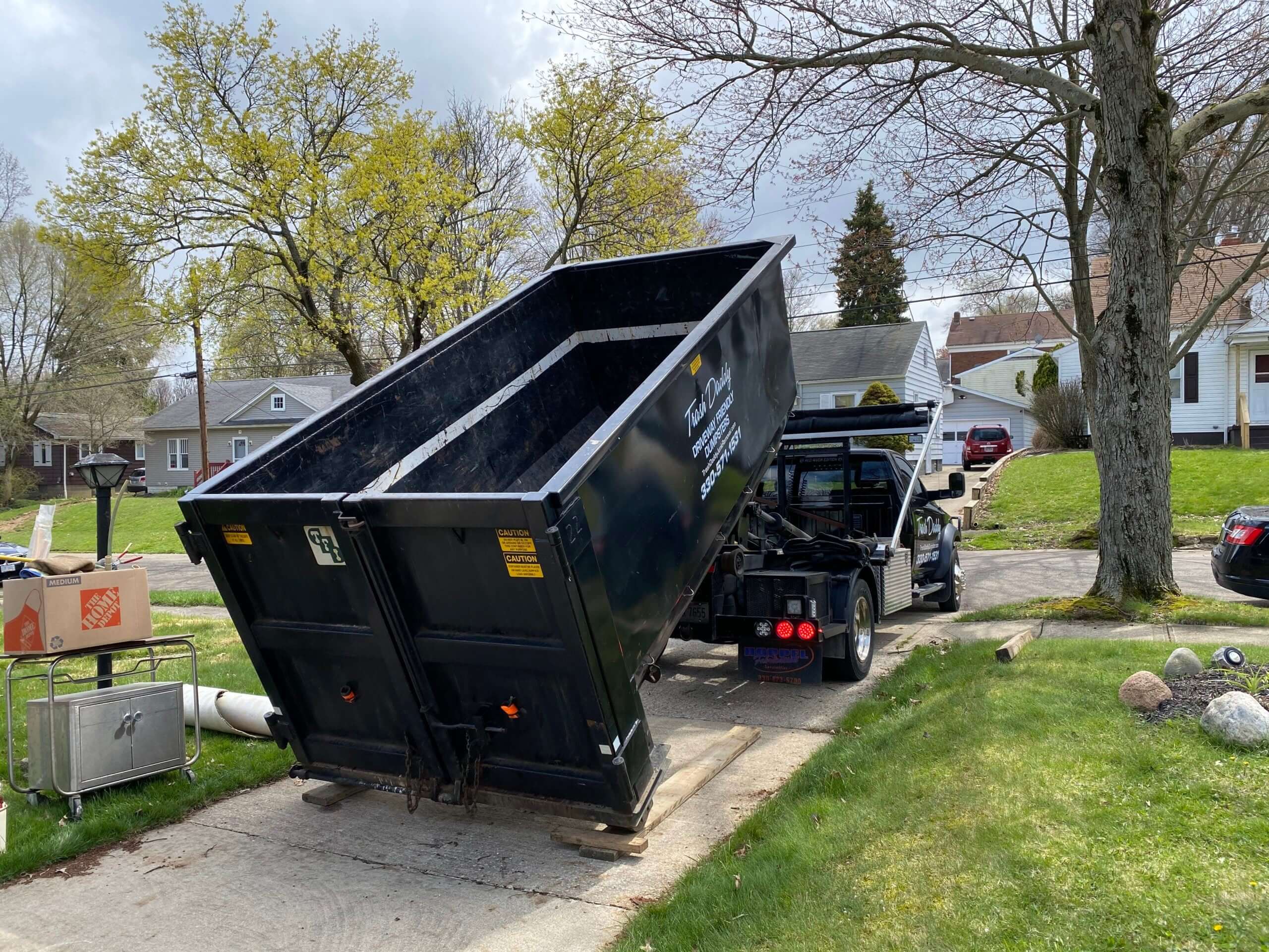 A black dumpster truck tilting to unload debris in a residential driveway under a cloudy sky.