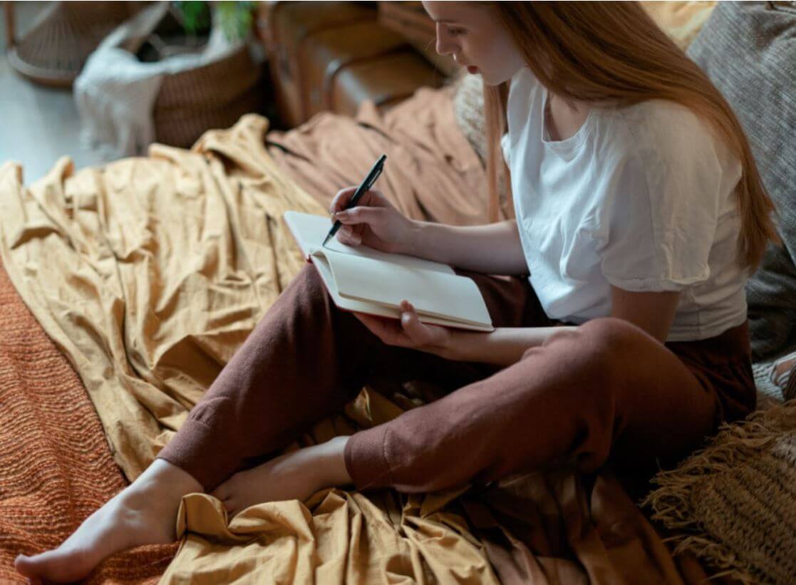 Woman sitting on a colorful blanket, writing in a notebook, showcasing a cozy and creative atmosphere.