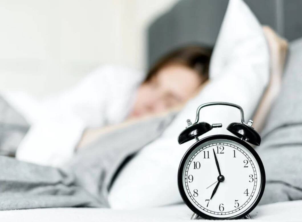 Alarm clock on bedside table with blurred person sleeping in background, symbolizing waking up or oversleeping.