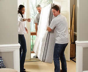 Two men carrying a mattress while a woman directs them at the front door of a home.