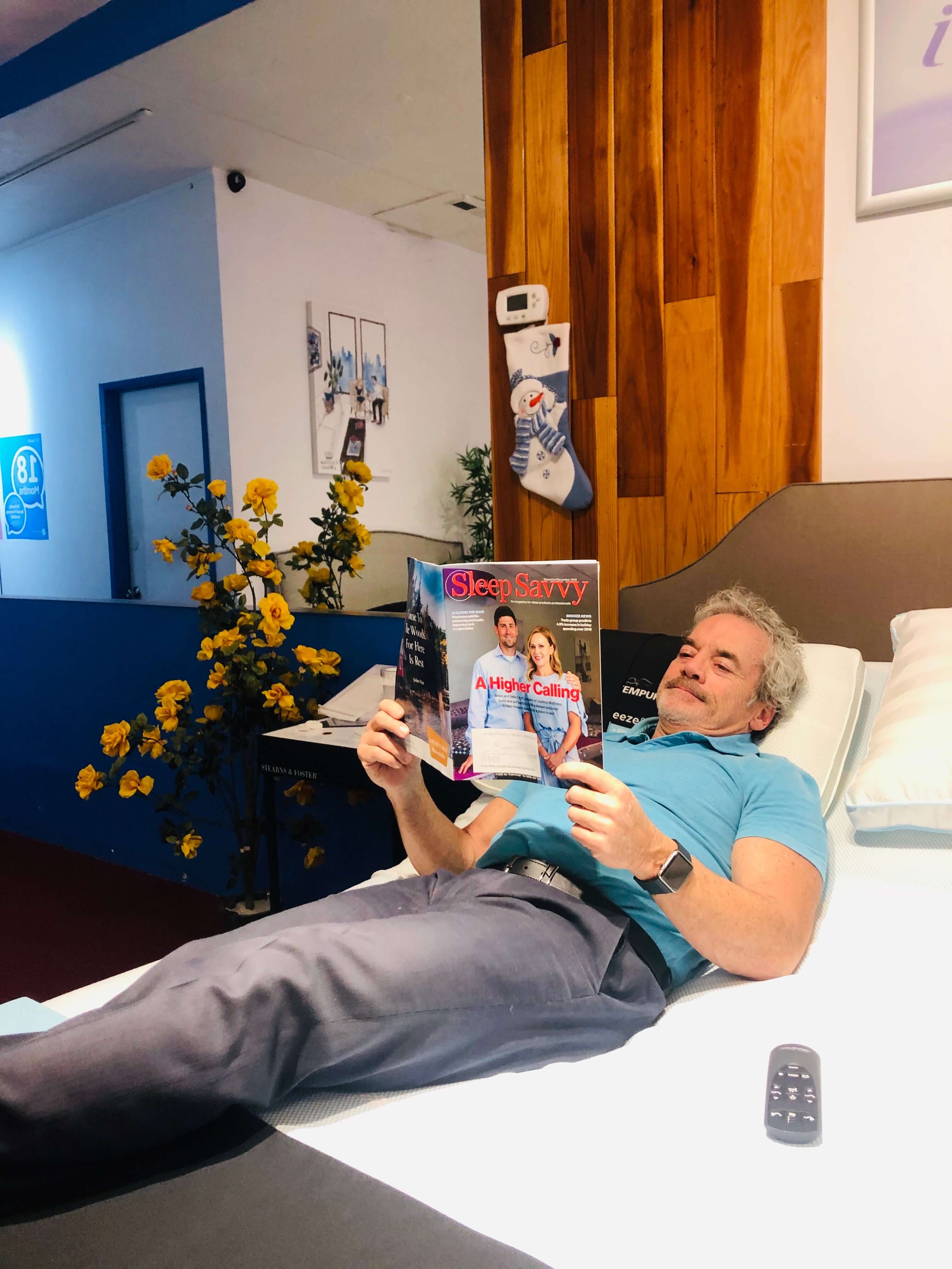 Man relaxing on a bed while reading Sleep Savvy magazine in a cozy room with floral decor.