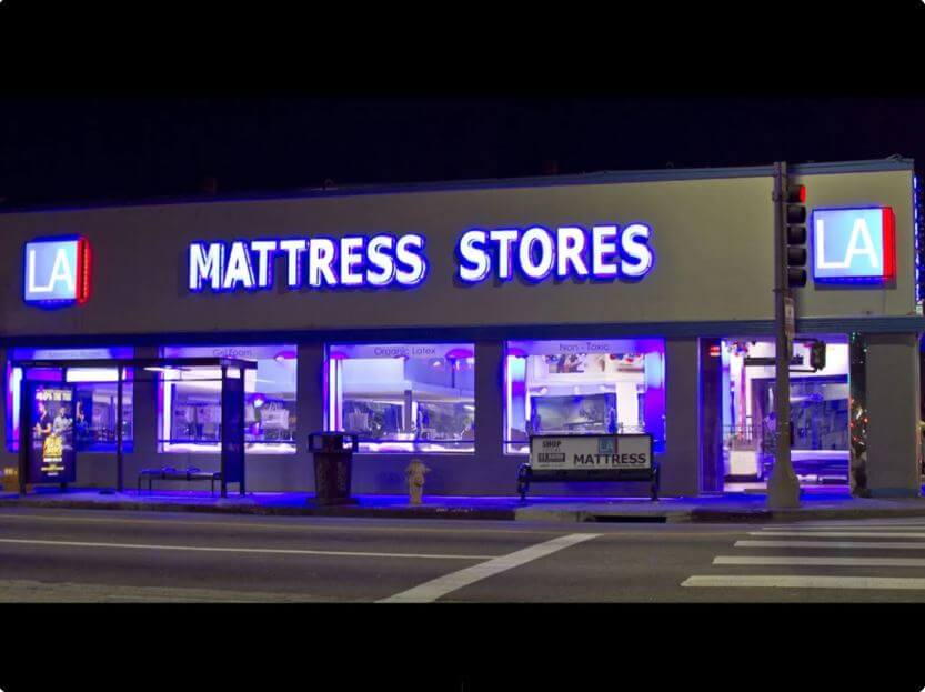 Brightly lit LA Mattress Stores at night, showcasing various mattress displays in the windows.