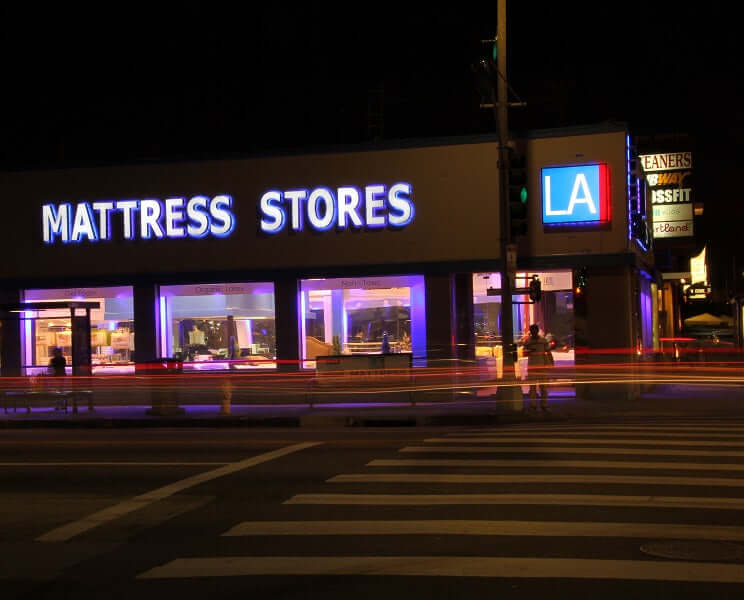 Brightly lit mattress store at night, showcasing a variety of mattresses with a busy urban street in Los Angeles.