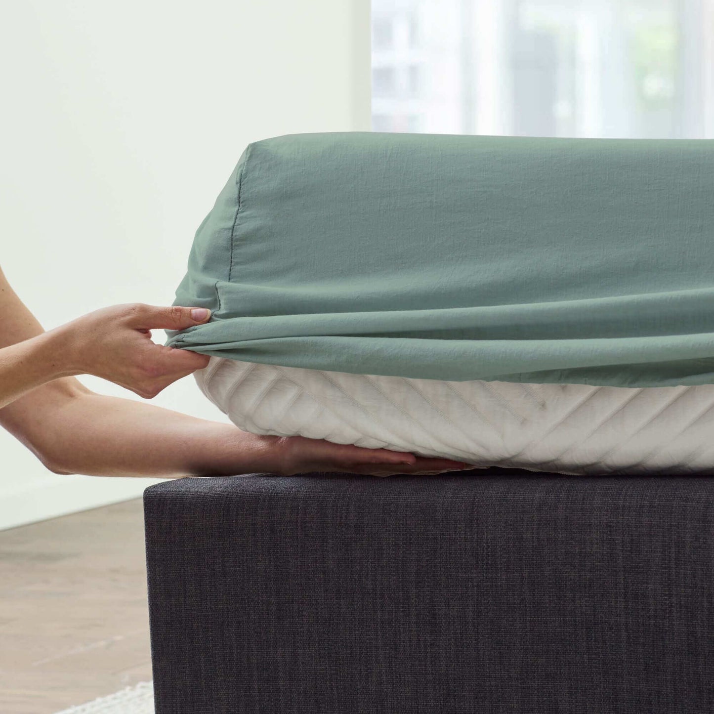 Person adjusting a light green sheet over a mattress, showcasing the softness and fit of Malouf Fog Linen Weave Cotton Sheet Set.