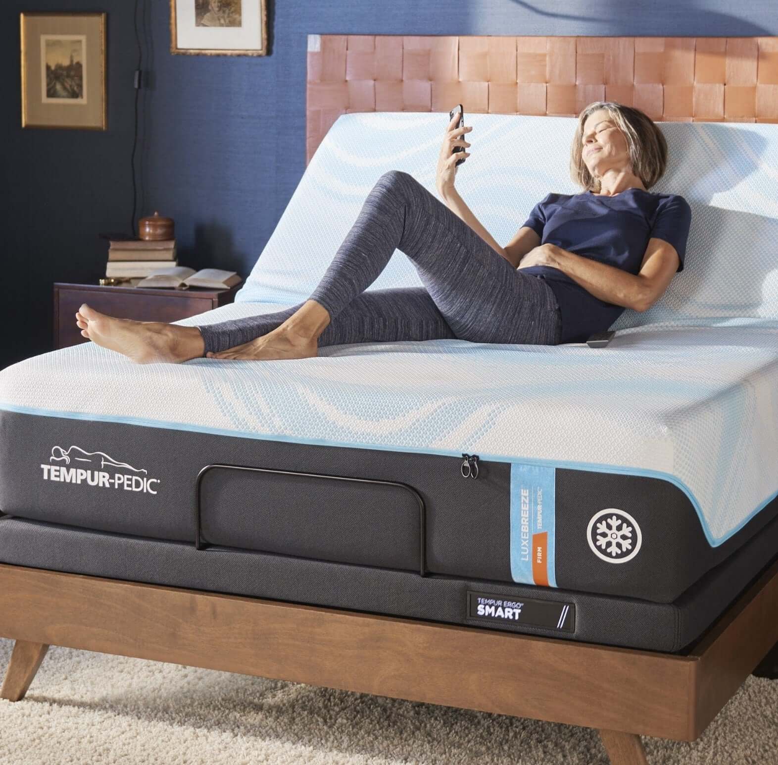 Woman relaxing on a Tempur-Pedic mattress, using her phone in a cozy bedroom setting.