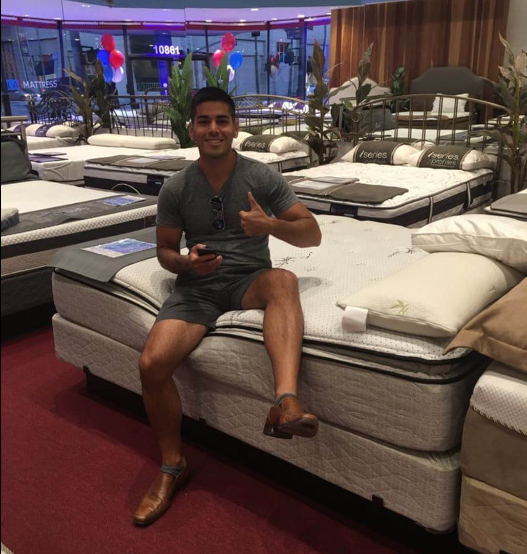 Young man smiling and giving a thumbs up while sitting on a mattress in a showroom with various beds displayed.