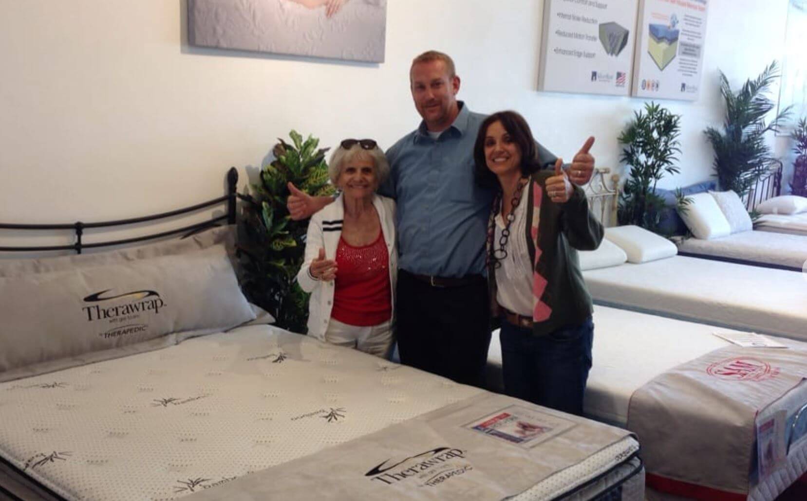 Group of three smiling individuals giving thumbs up in a mattress showroom, showcasing a Therawrap mattress.