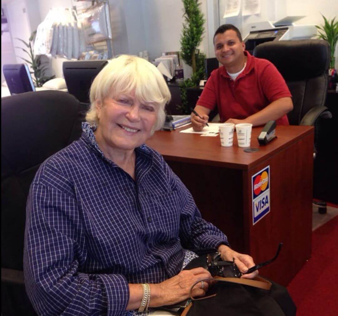 Smiling older woman in a blue shirt sitting in an office, with a man at a desk in the background, showcasing a friendly atmosphere.