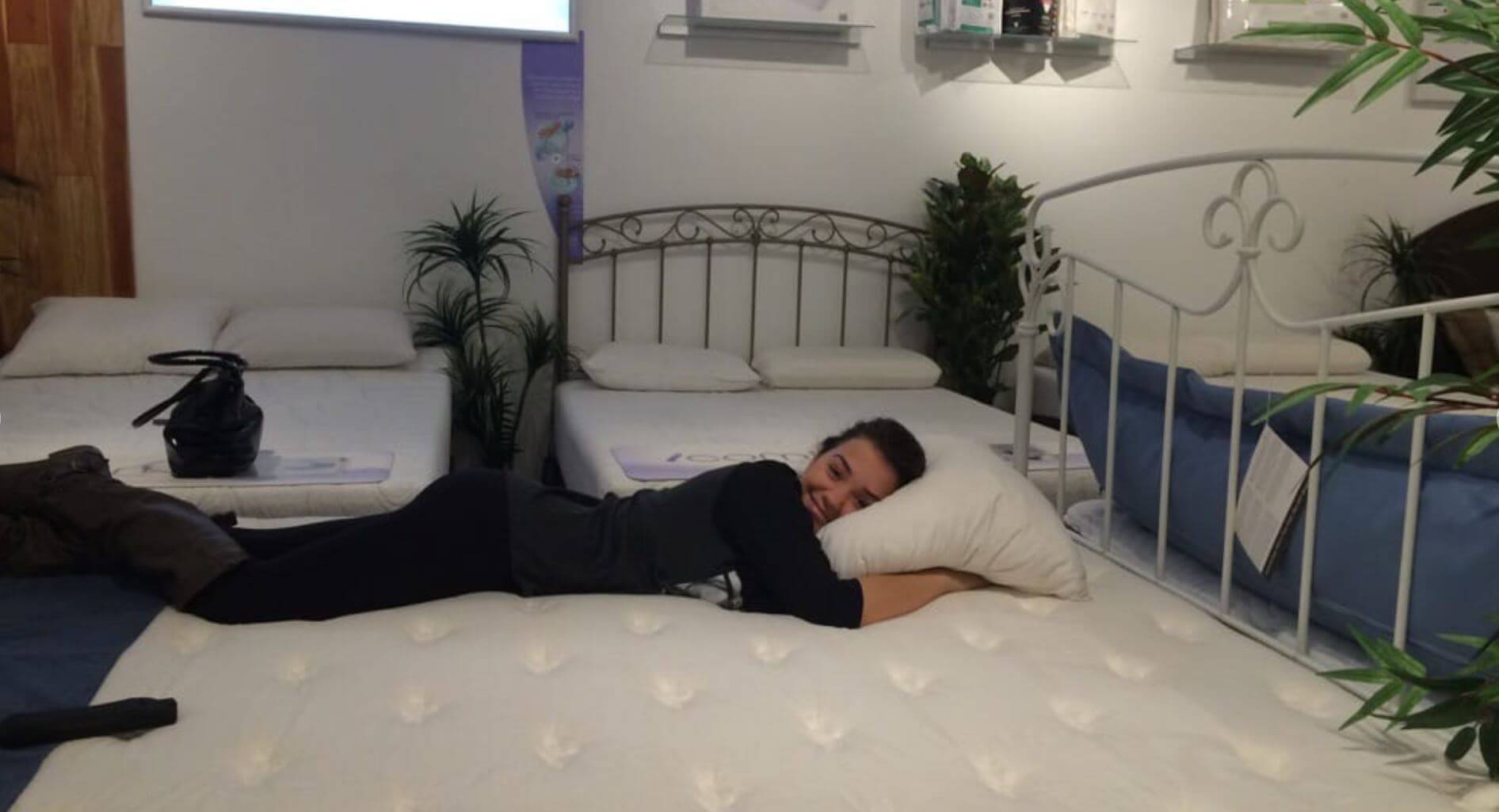 Woman testing a comfortable mattress while smiling in a home furnishings store, surrounded by other bedding options.