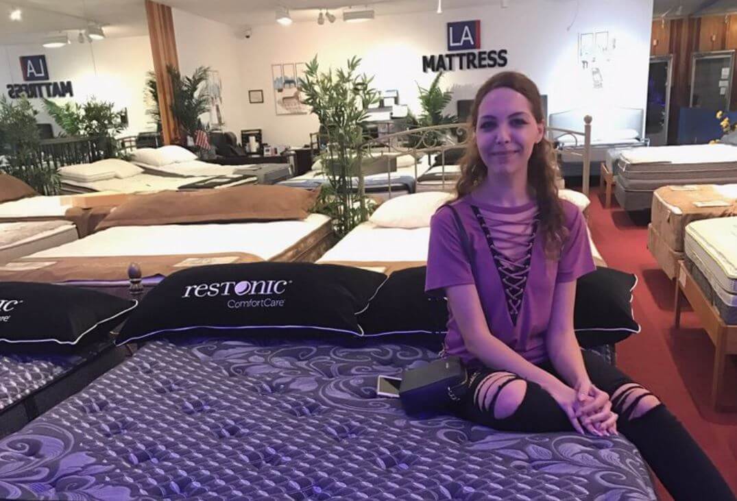 Woman sitting on a Restonic mattress in a mattress showroom with various beds visible in the background.