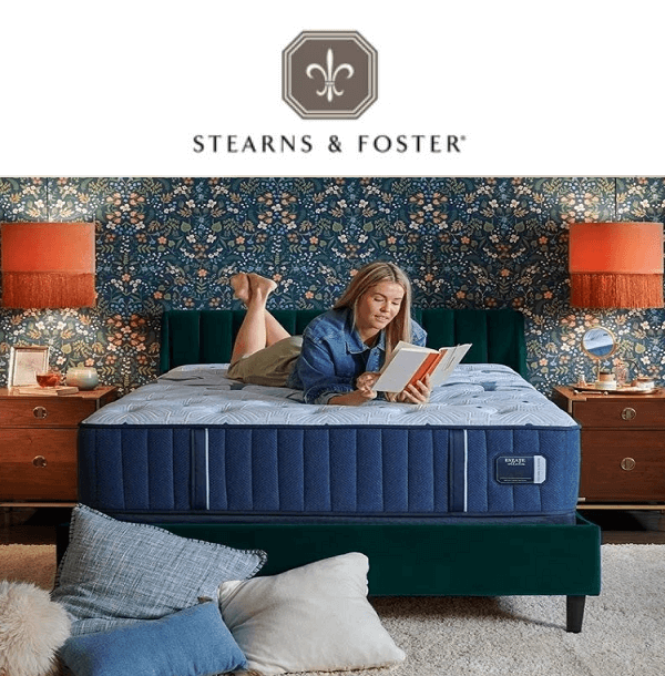 Woman reading on a Stearns & Foster mattress in a stylish bedroom setting with floral wallpaper and cozy decor.
