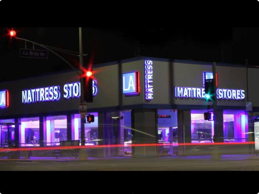 Night view of LA Mattress Stores featuring illuminated signage and vibrant colors on a busy intersection.