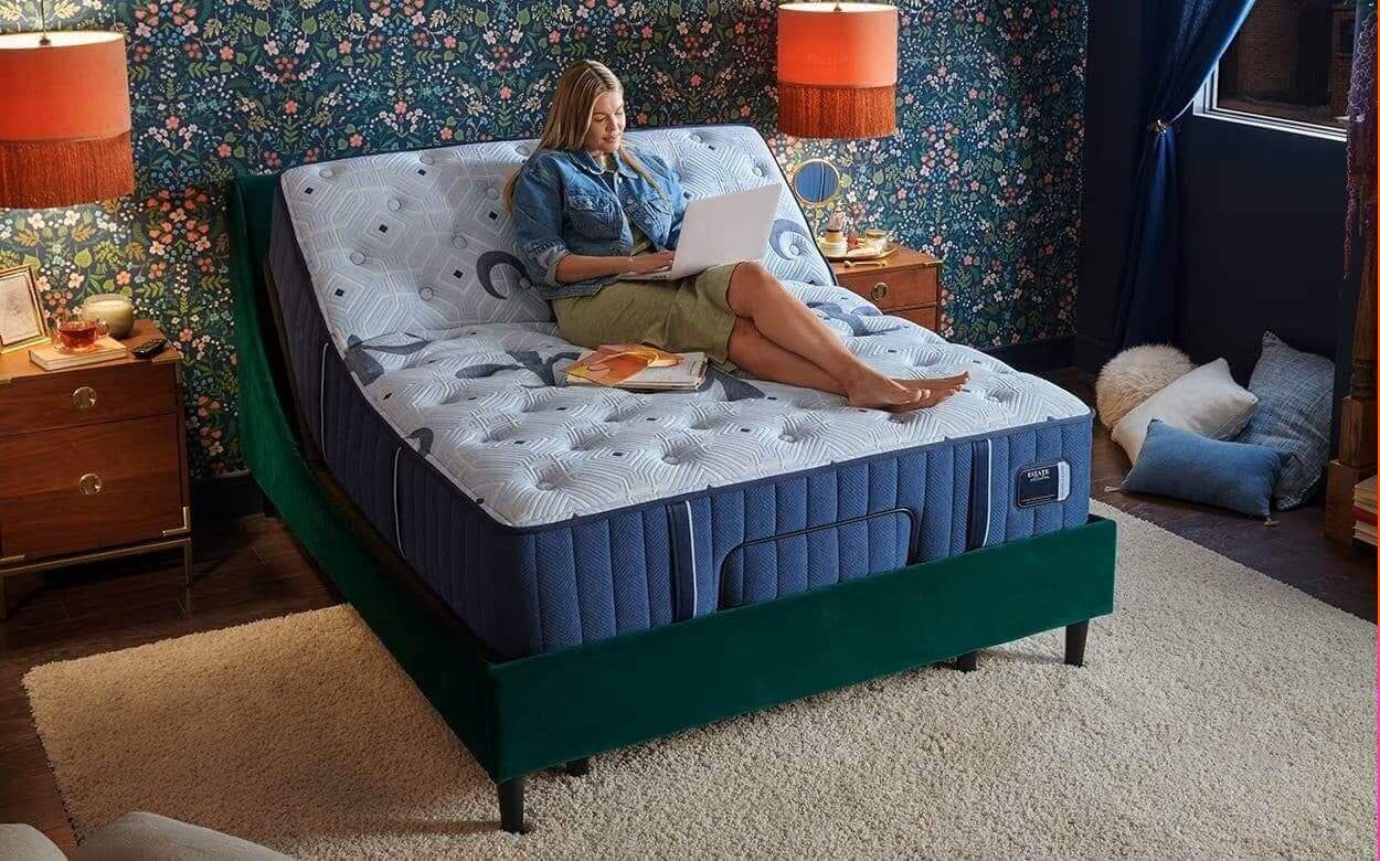 Woman using a laptop while sitting on a Stearns & Foster Estate 14.5" Plush Mattress in a stylish bedroom setting.