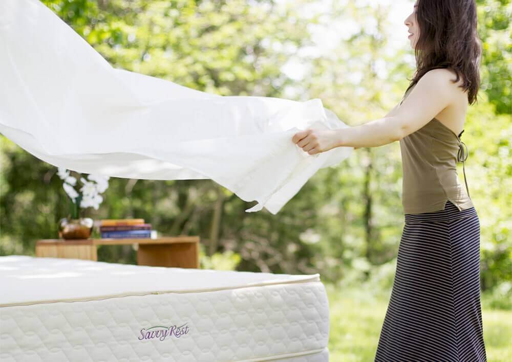 Woman elegantly draping Savvy Rest organic cotton sheet over a mattress outdoors in a natural setting.