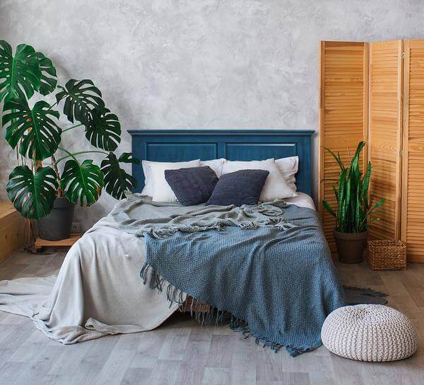 Cozy bedroom with a blue headboard, lush plants, and a textured blanket, perfect for relaxation and comfort.