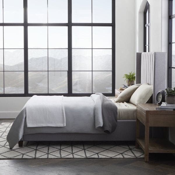 Cozy bedroom with a modern bed dressed in soft gray sheets and blankets, featuring large windows and a stylish wooden nightstand.