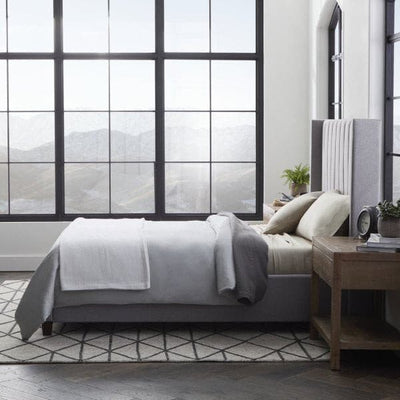 Cozy bedroom with a modern bed dressed in soft gray sheets and blankets, featuring large windows and a stylish wooden nightstand.