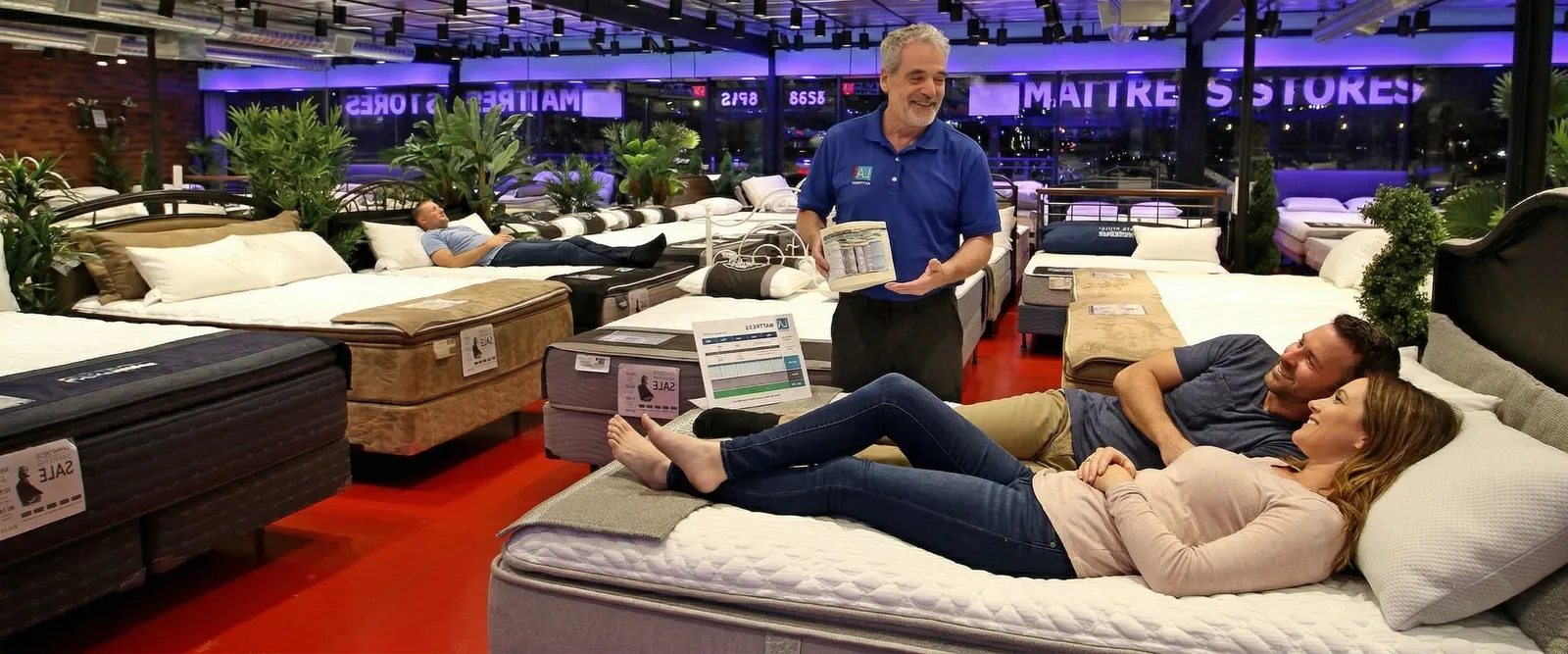 A couple lying on a mattress while an LA Mattress Store consultant helps them choose, inside a Los Angeles showroom