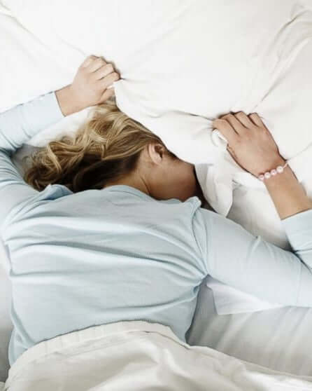 Woman buried under a pillow in bed, expressing frustration or exhaustion.