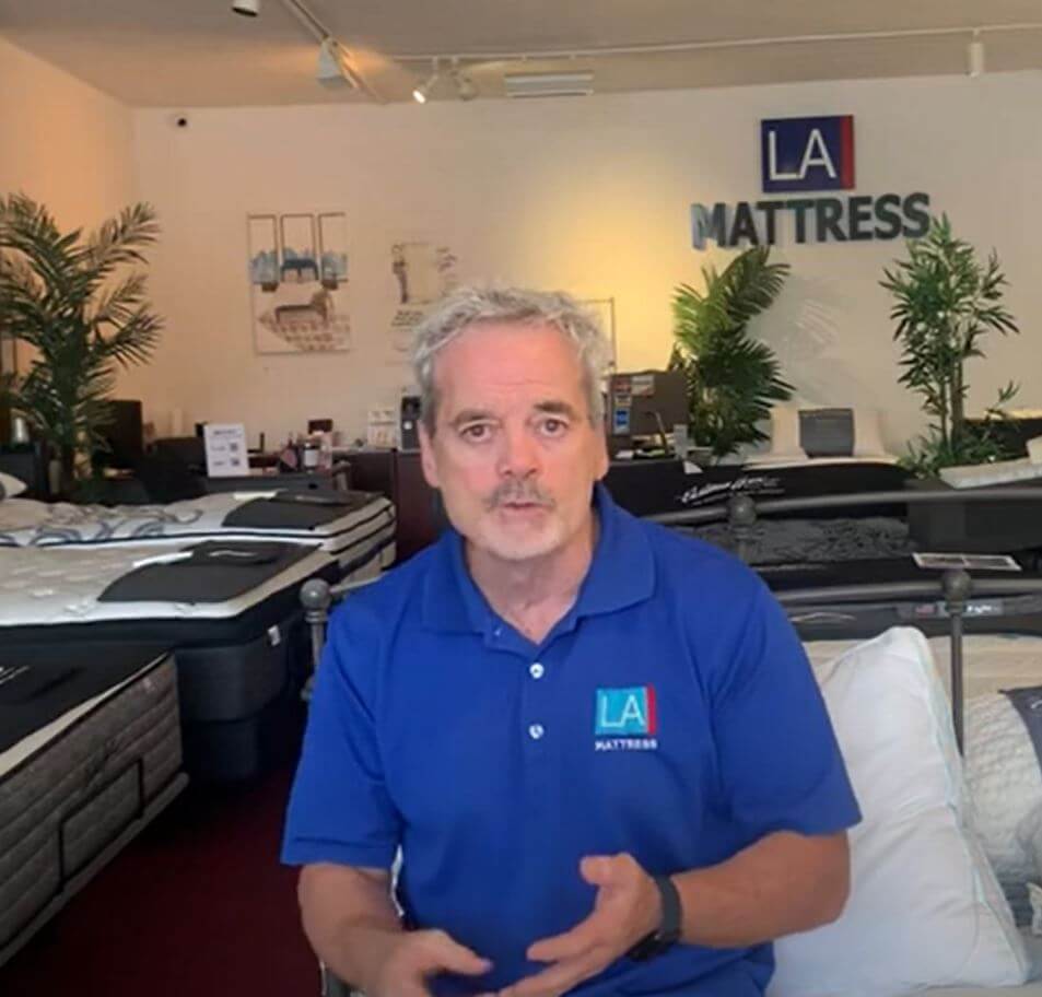 Man in blue shirt discussing mattresses in an LA Mattress showroom with plants and beds in the background.