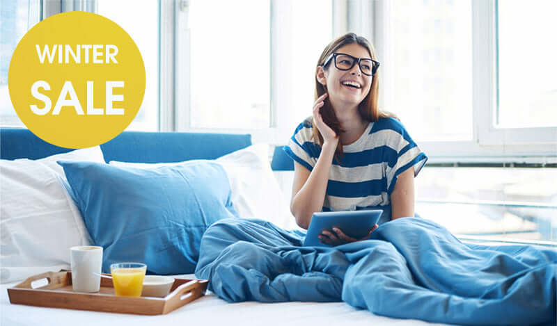 Young woman in striped shirt enjoying breakfast in bed during winter sale, smiling while holding a tablet.