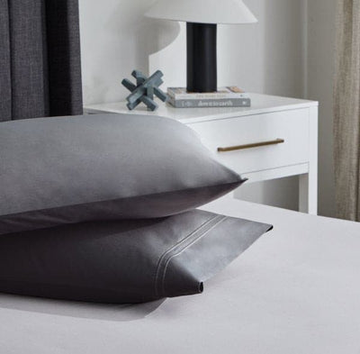 Gray Supima® cotton pillows stacked on a bed beside a nightstand with a lamp and decorative items.